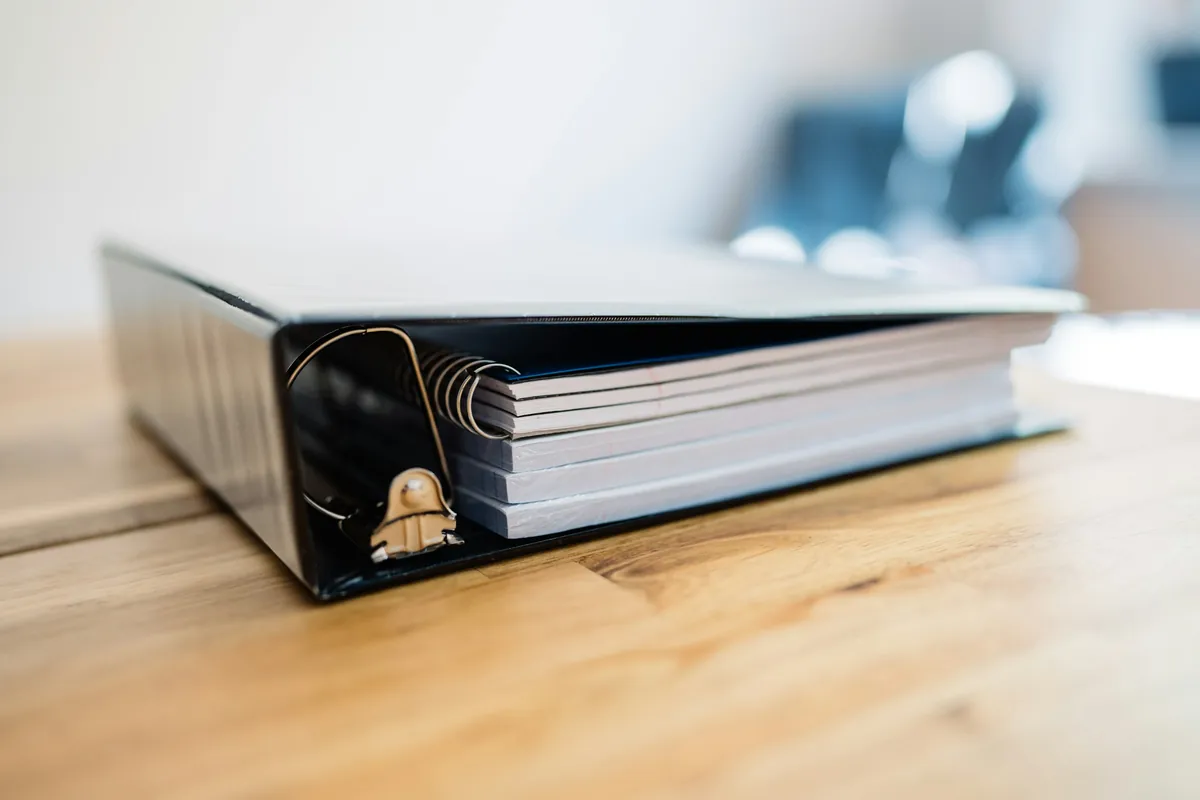 A thick black three-ring binder full of papers, lying on a wooden desk.